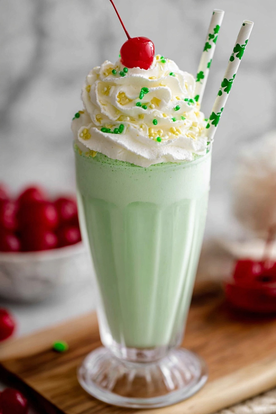 A tall clear glass holds a light green milkshake with a creamy, smooth texture, topped with a thick swirl of white whipped cream sprinkled with tiny green and yellow sugar shapes. A bright red cherry sits on top of the whipped cream, and two white straws with green shamrock patterns stick out from one side. The glass stands on a wooden board with a white marbled background, and a bowl of red cherries can be seen blurred in the back. photo taken with an iphone --ar 2:3 --v 7