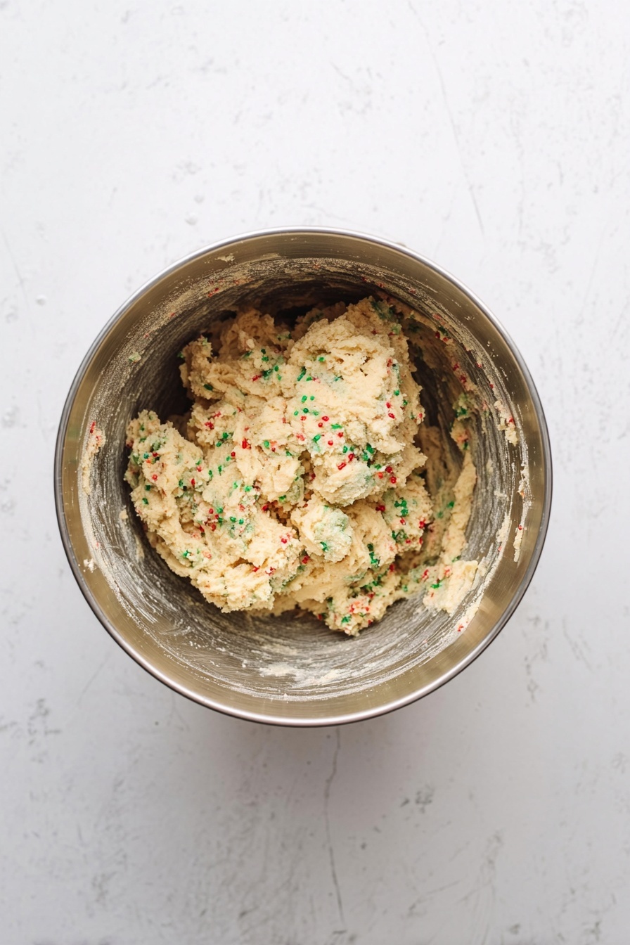 A top-down view of a metal mixing bowl filled halfway with creamy cookie dough that has green and red sprinkles mixed throughout. The dough has a slightly rough texture with specks of flour on some parts. The bowl sits on a white marbled surface with soft, natural lighting highlighting the texture of the dough and the shiny metal of the bowl. photo taken with an iphone --ar 2:3 --v 7