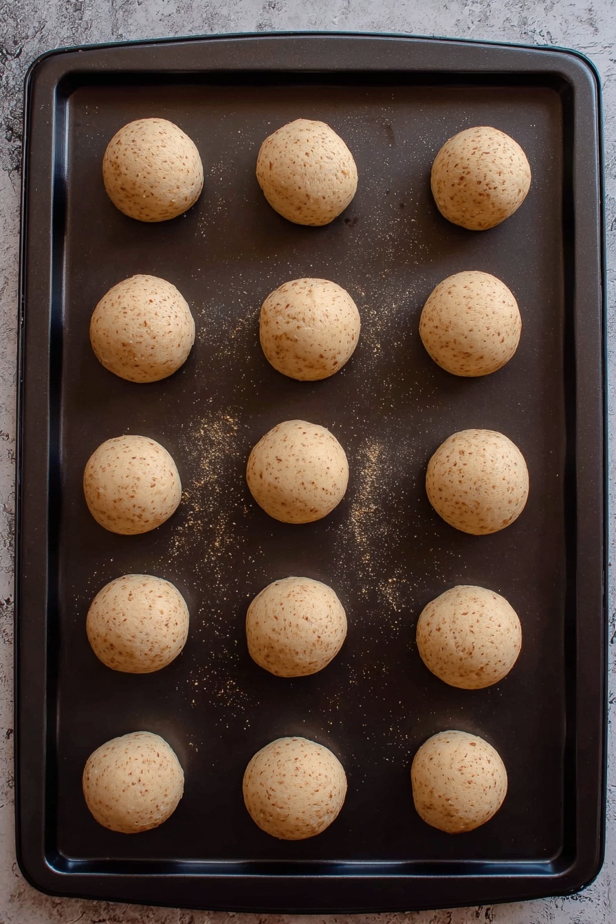 A round open tin with a dark blue and green plaid pattern and white floral decorations is filled with three layers of round cookies, each covered in white powdered sugar with small red and green sprinkles on top. The cookies are stacked loosely with some overlapping and resting on a white parchment paper lining inside the tin. The tin is placed on a smooth white marbled surface, surrounded by white sheer ribbon with stripes and shiny silver ornaments. In the background, the tin’s lid is propped up showing the same patterned design, and soft white fabric adds a delicate touch. Photo taken with an iphone --ar 2:3 --v 7