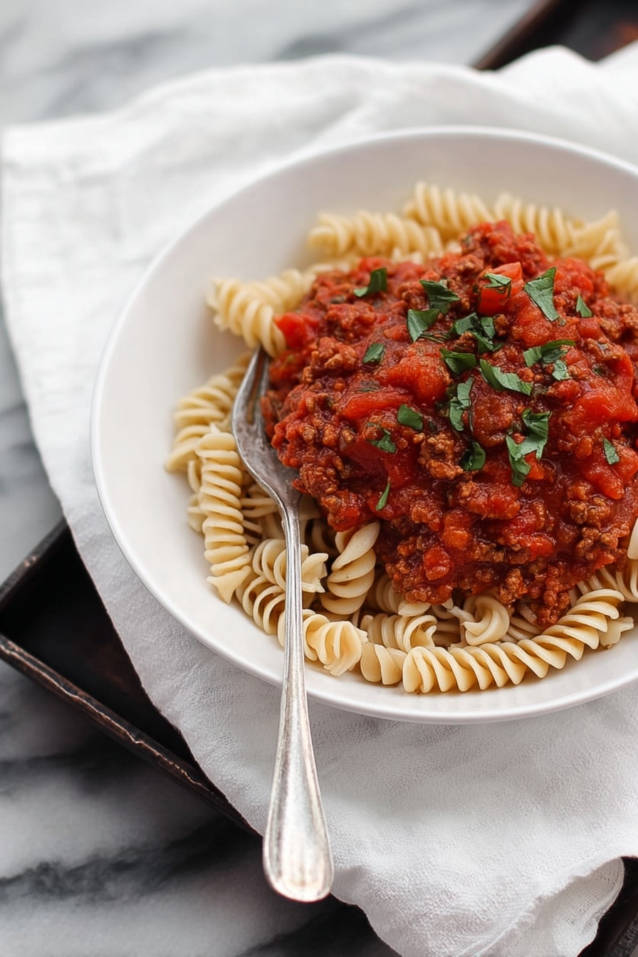 A white bowl filled with a spiraled pasta base, light beige in color with a smooth texture, topped with a thick red tomato sauce containing small pieces of cooked brown meat and finely chopped onions, sprinkled with green herb leaves. The bowl is placed on a white cloth on a metal tray with a rustic look, accompanied by a small white bowl of dark red olives and a clear glass of water. A fork and knife are set next to the bowl on the cloth, with a green basil leaf beside it on the tray. The background is a white marbled texture. photo taken with an iphone --ar 2:3 --v 7